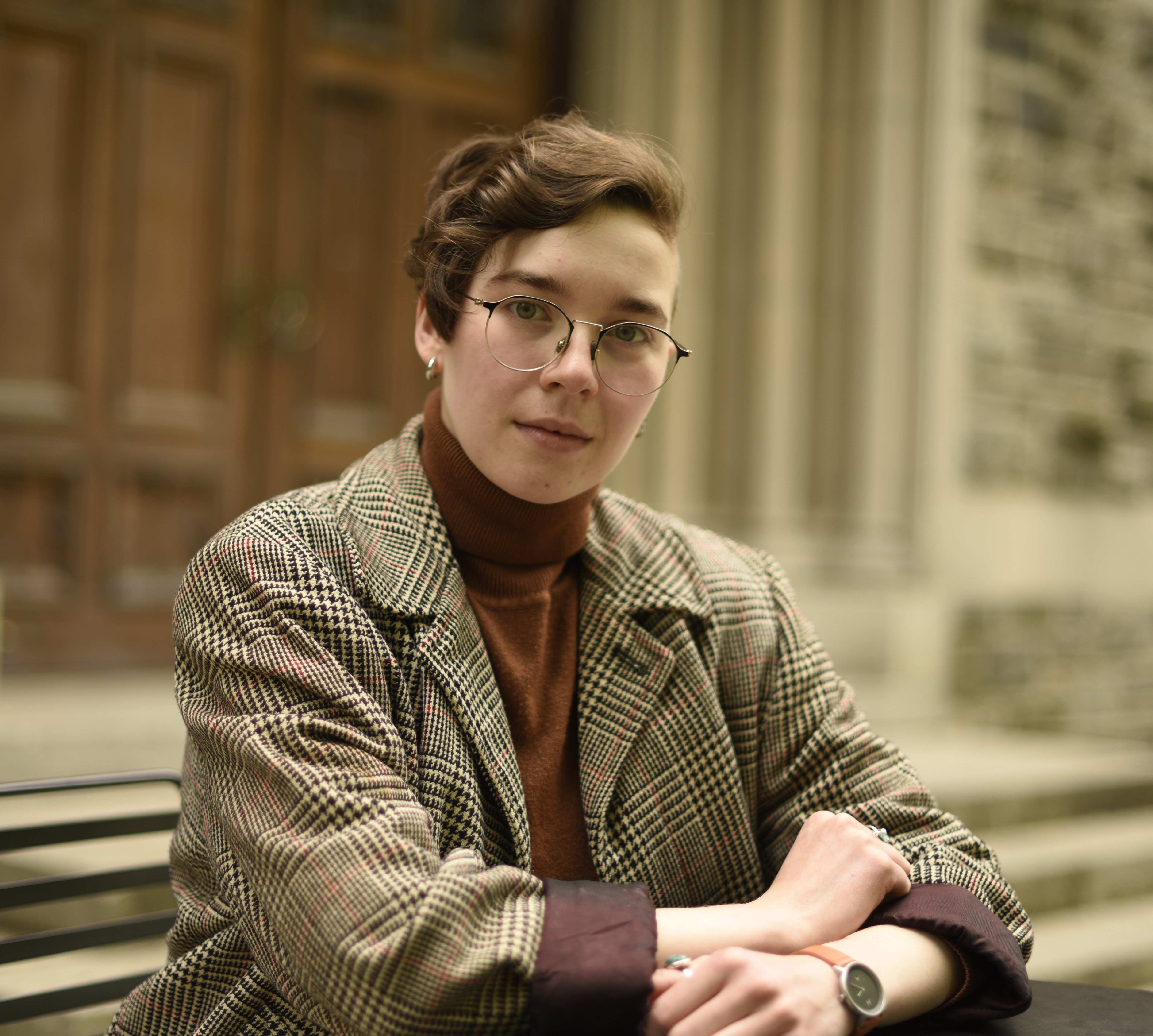 A white person with brown hair, long on one side and short on the other, wearing a brown turtleneck and sport coat, sits in front of a wooden door. They are not smiling, nor are they frowning.